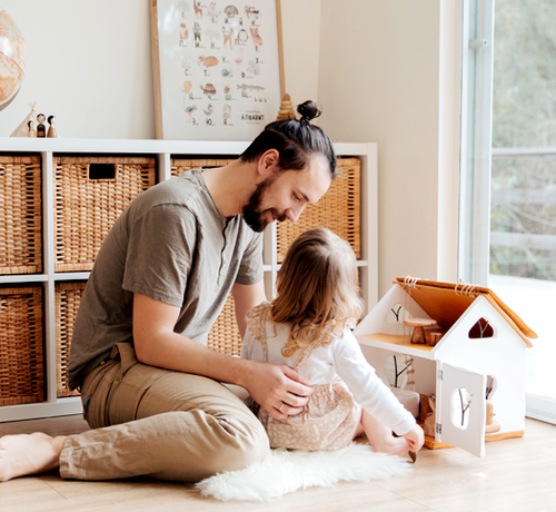 Dad sitting on the floor at his toddler’s level, playing with her and a wooden dollhouse in a bright living room - Mustela Australia - 1