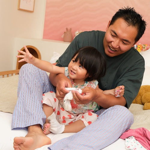 Father sitting on the floor at home, smiling and playing with his toddler daughter as he gently holds her hands, creating a warm and joyful family moment. - Mustela Australia - 1